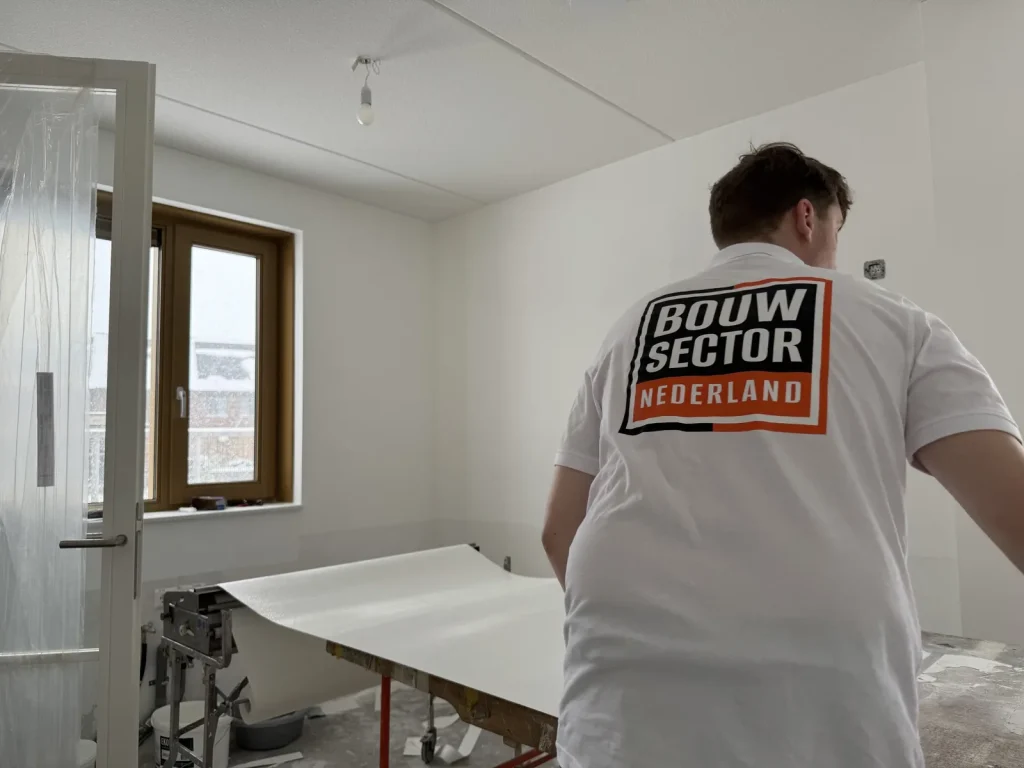 Behanger van Bouwsector Nederland in een wit poloshirt legt een baan glasvlies op de plaktafel in een nieuwbouwkamer