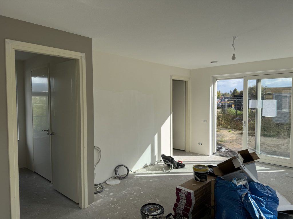 Woonkamer van een net opgeleverde nieuwbouwwoning met kale wanden, een betonvloer, verfemmers en dozen voor de tuindeur