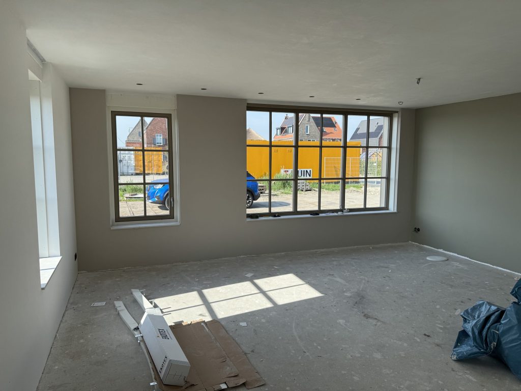 Ruime nieuwbouwkamer met wanden in een warme grijstint en grote ramen met zicht op nieuwe huizen