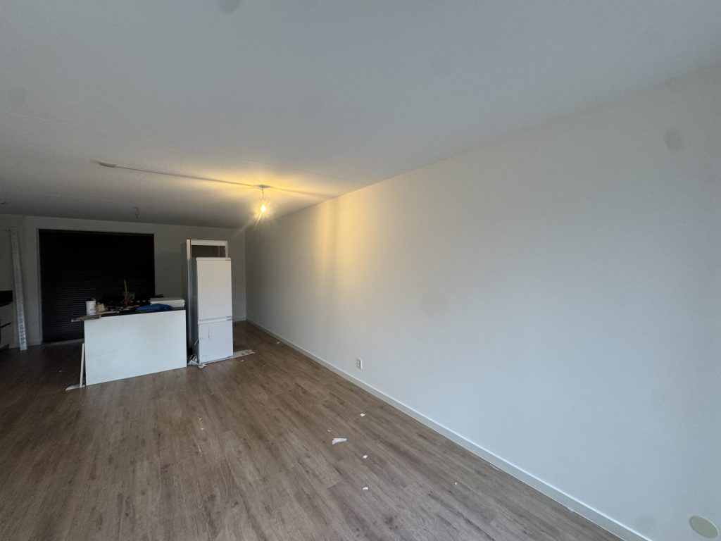 Woonkamer van een nieuw appartement met een houten vloer, gladde witte wanden en een open keuken achterin