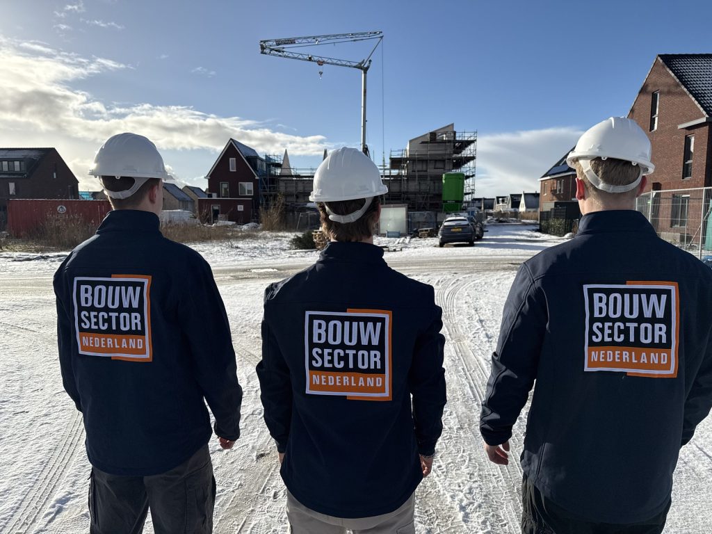 Drie vakmensen van Bouwsector Nederland met witte helmen en donkerblauwe jassen met logo op een besneeuwde nieuwbouwlocatie met een bouwkraan
